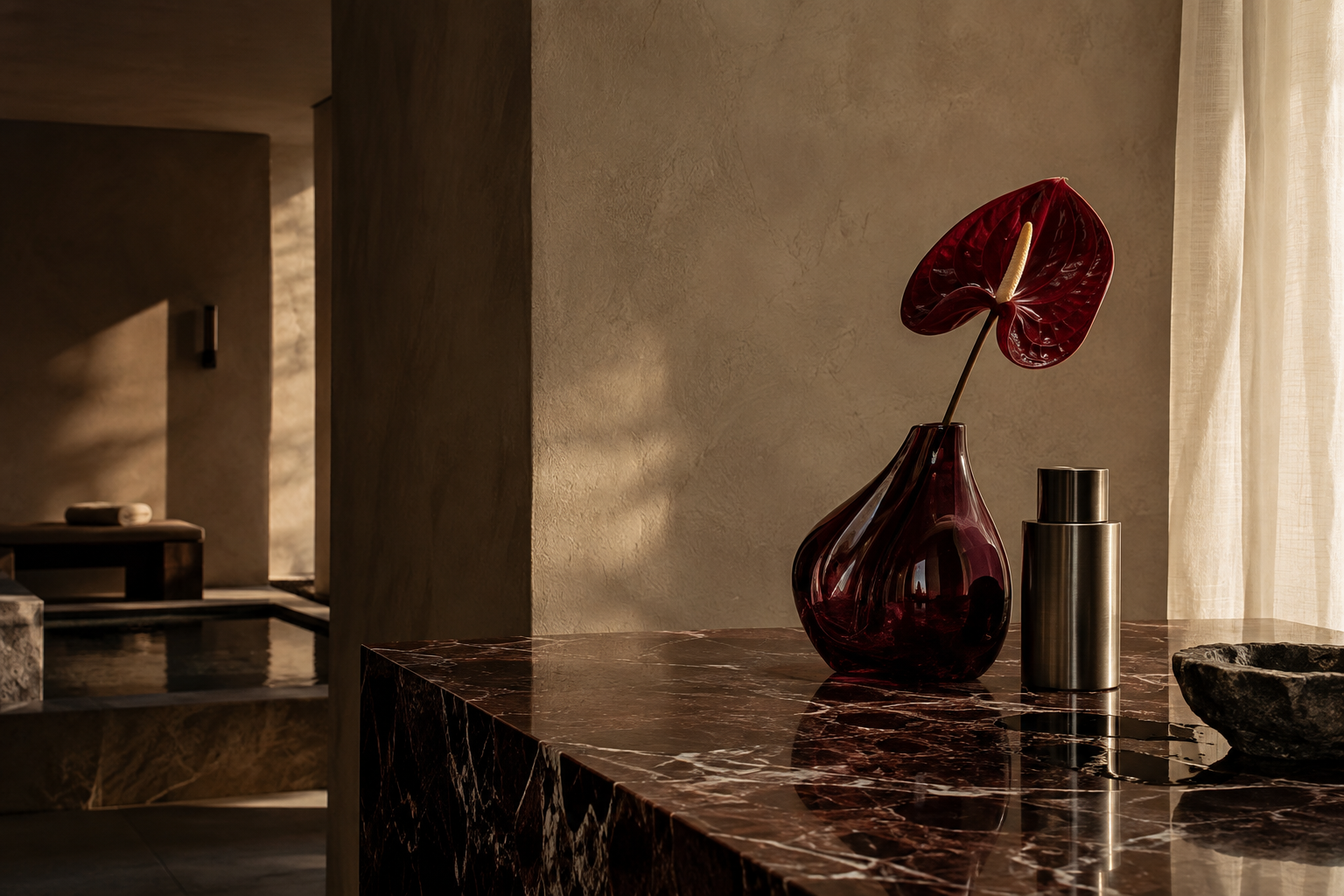 Burgundy anthurium, sculptural glass and a metal vessel in a sunlit stone interior.