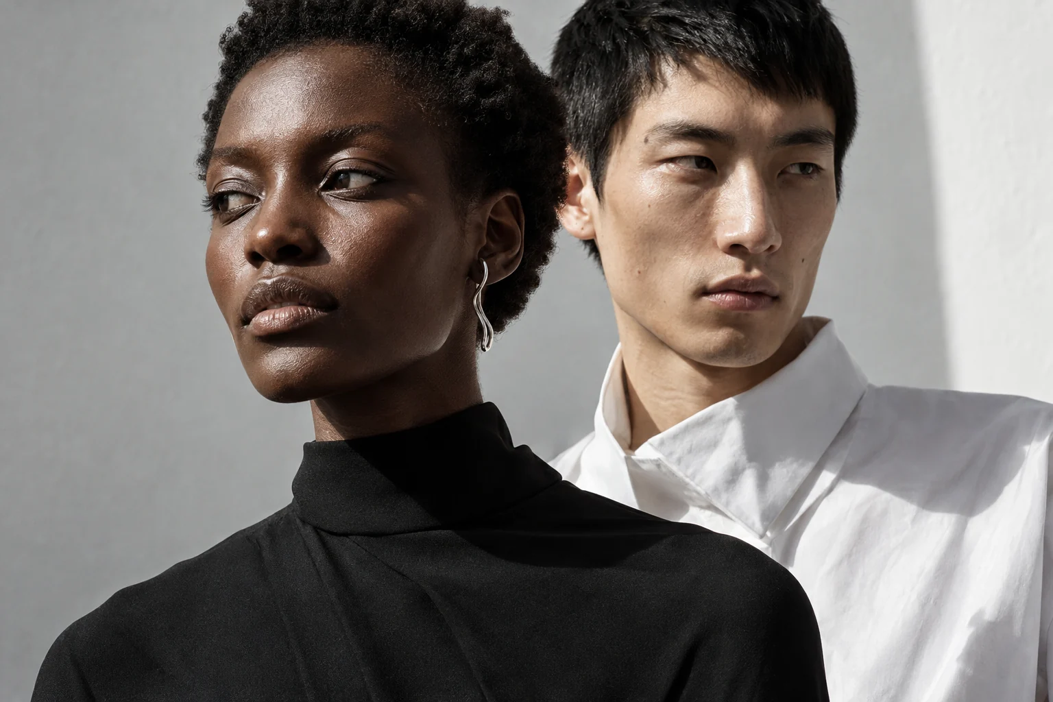 Two adult fashion models in sculptural black and white tailoring, photographed in hard studio light.