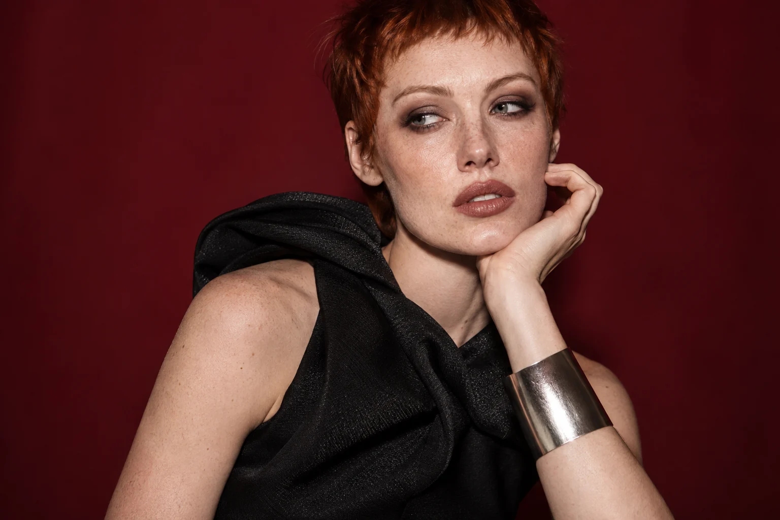 An adult model with cropped copper hair, black eveningwear and a silver cuff against an oxblood backdrop.