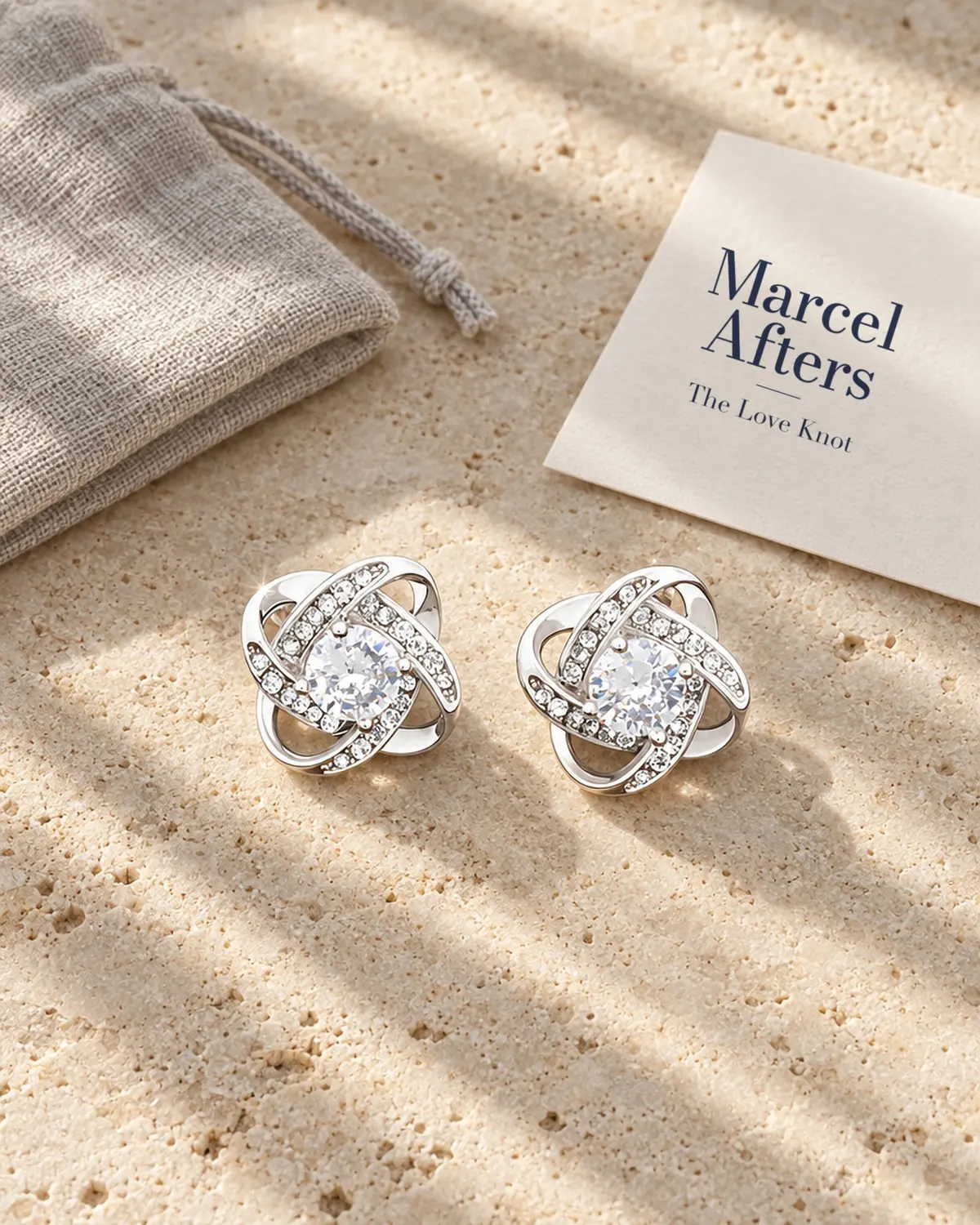 Love-knot crystal stud earrings beside a Marcel Afters branded card on limestone.