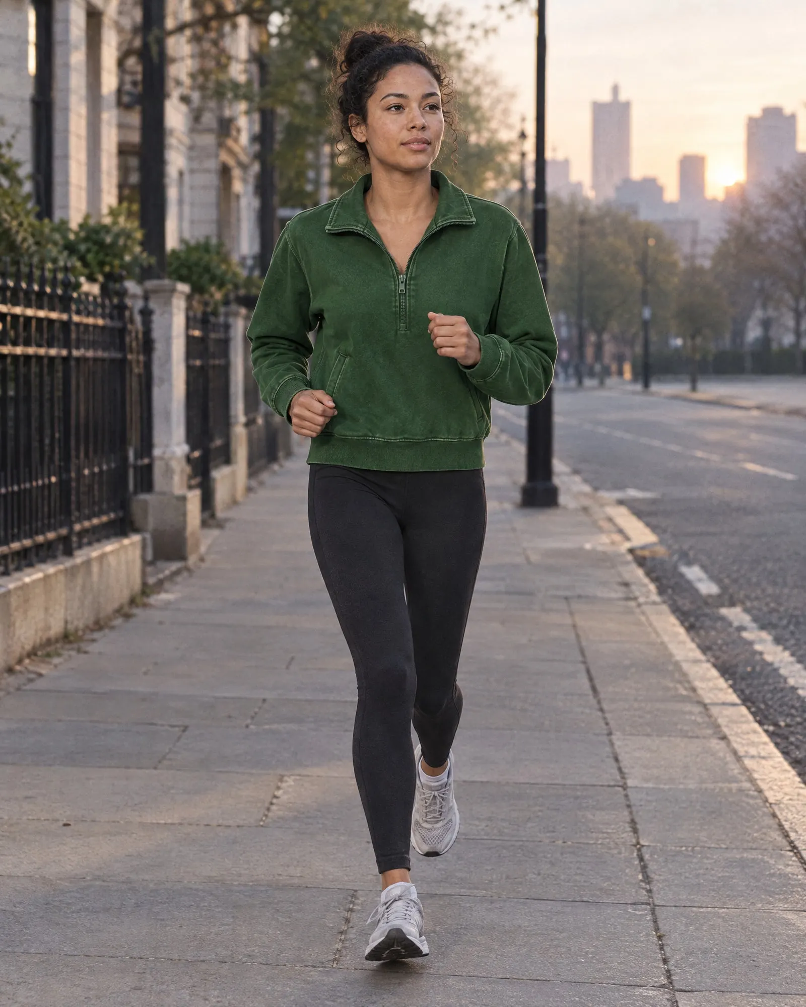A woman runs down a city street at dawn in a green washed quarter-zip and black leggings.