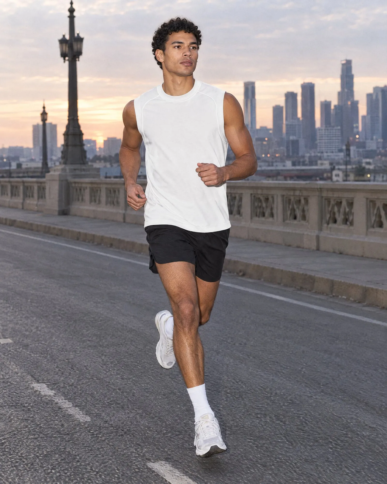 A man runs across a bridge at sunrise in a white performance tank and black shorts.