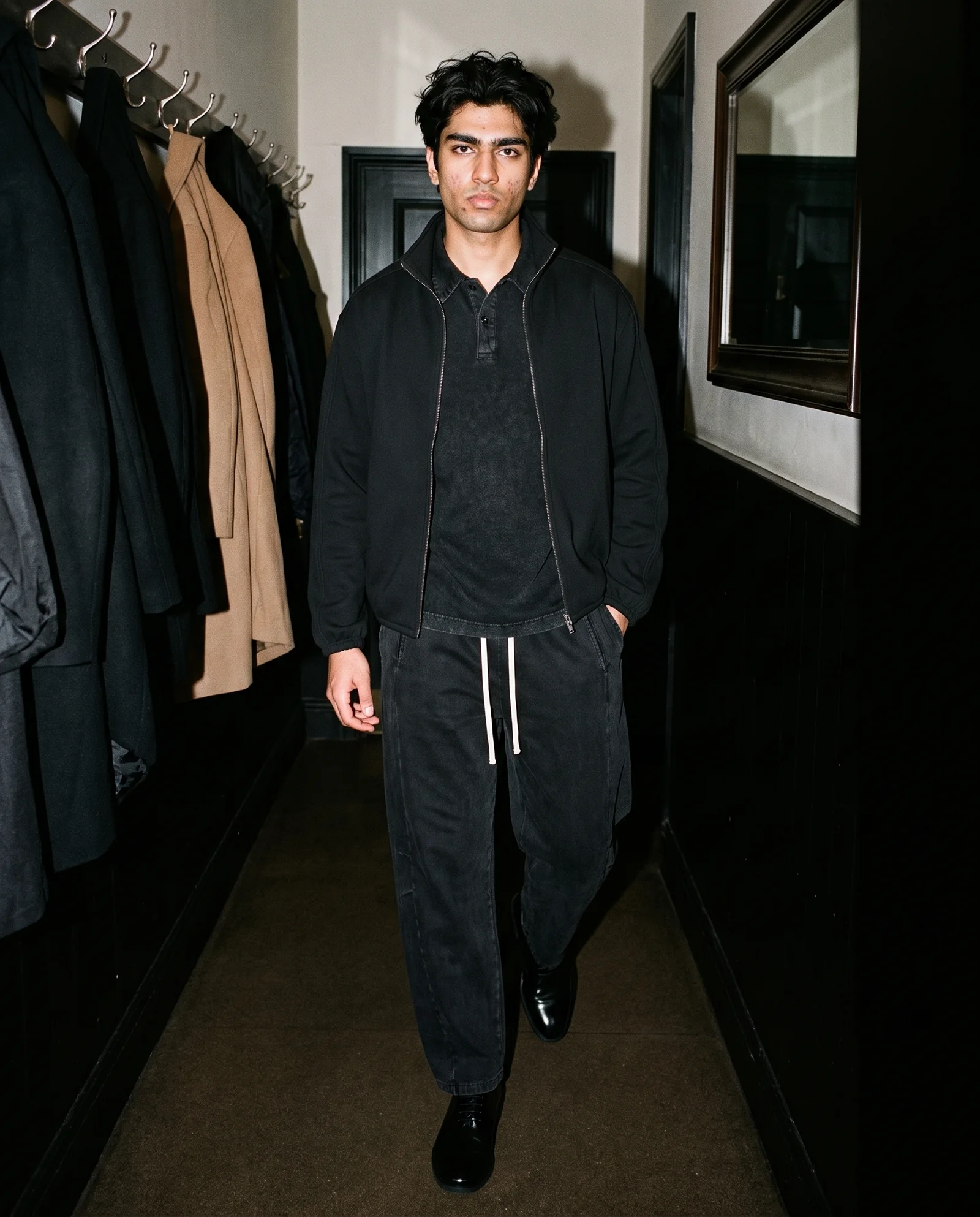 A man in a black jacket, black shirt, black pants, and black shoes, standing in a hallway.