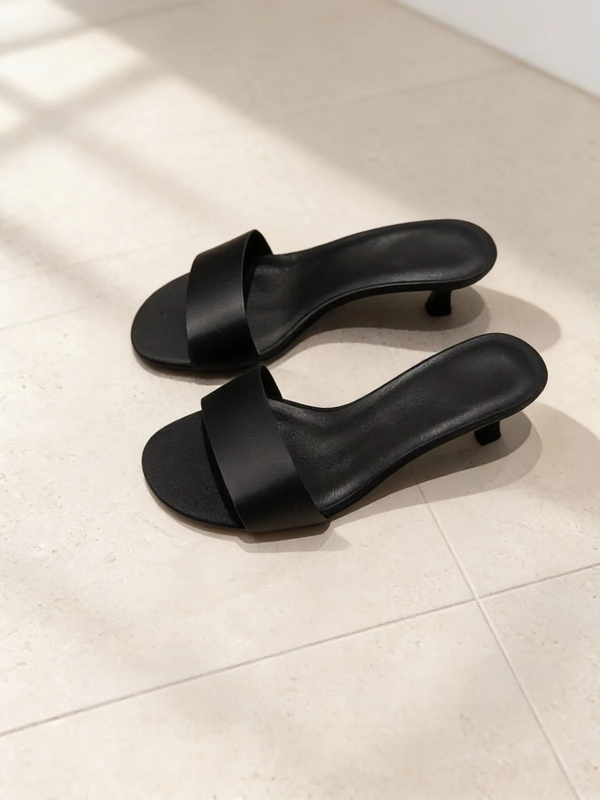 The Room Service Slide in black, a pair of low-heel slides on limestone.
